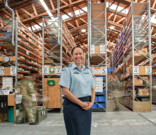 A woman stands in a warehouse with her hands together in front of her, she is wearing a RNZAF uniform. In the background, people in uniform are blurred by motion as industrial shelving, nets, trays and boxes. are lit by skylights in the ceiling.
